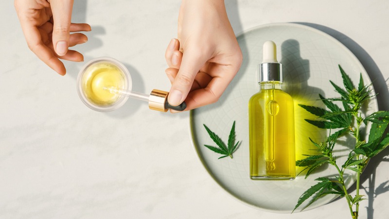 Female Hands Holding CBD Oil Dropper With CBD Oil Bottle and Hemp Leaves on a Table