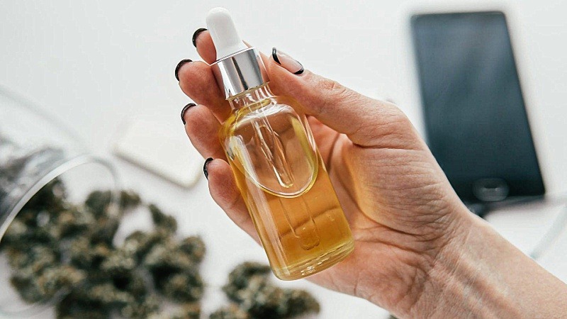 Hand holding a bottle of CBD oil extract with dried hemp flowers and a phone at the back